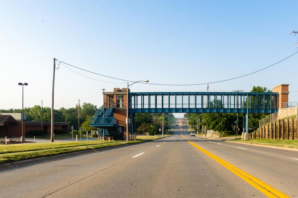 Wide road with a yellow center line passing under a pedestrian bridge, with trees and utility poles along the side.