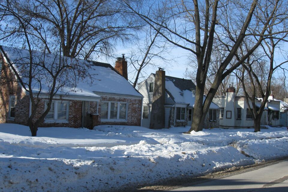 Snow-covered yards and roofs on a row of small houses along a residential street under bare trees and a blue sky.
