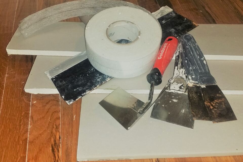 Cut pieces of drywall on a wood floor with a roll of drywall tape and several taping and putty knives.