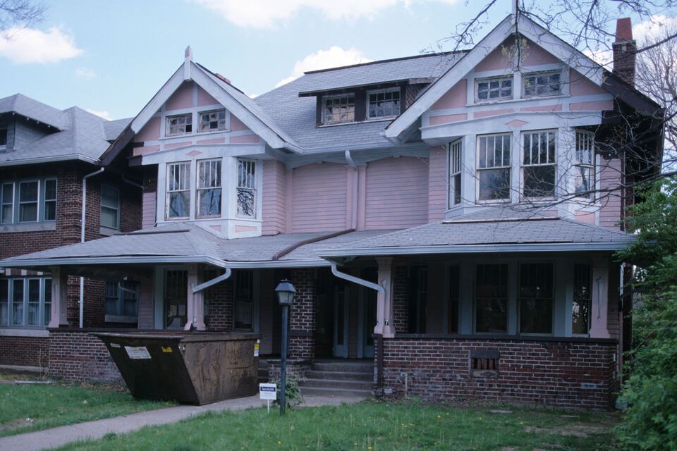 Large two-story early-1900s house with a wide front porch and a roll-off dumpster sitting on the front lawn.